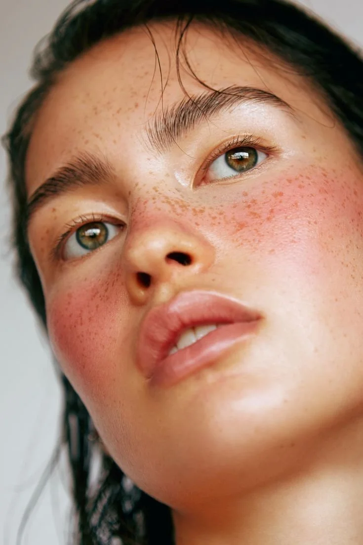 Close-up of a woman's face, showing her light brown eyes, dark wet hair, and flushed cheeks with freckles.